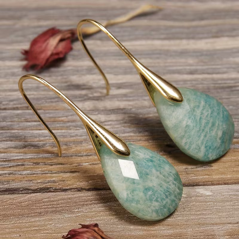 Faceted aqua-green teardrop earrings with long tapered gold-tone hooks on wood.