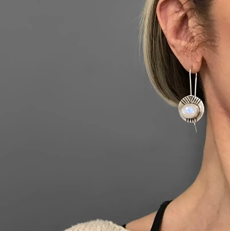 Model wearing a round pale iridescent earring in a silver-tone sunburst frame.