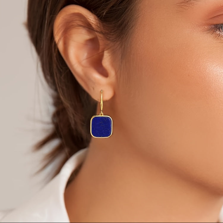 Model wearing a dark-blue rounded square earring in a gold-tone frame.