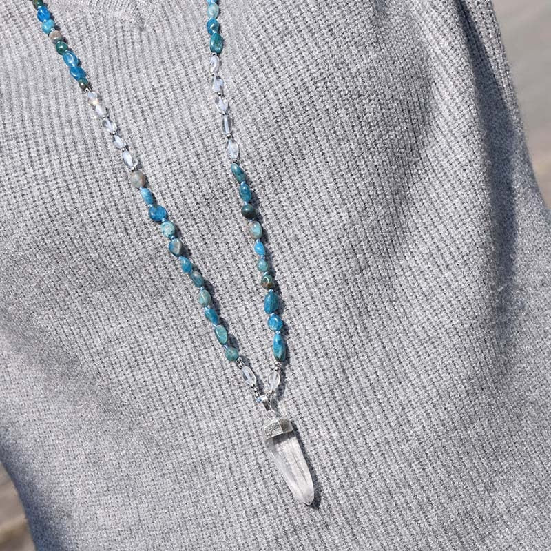 Model wearing a long blue beaded necklace with clear accents and a clear pointed pendant over a gray knit.