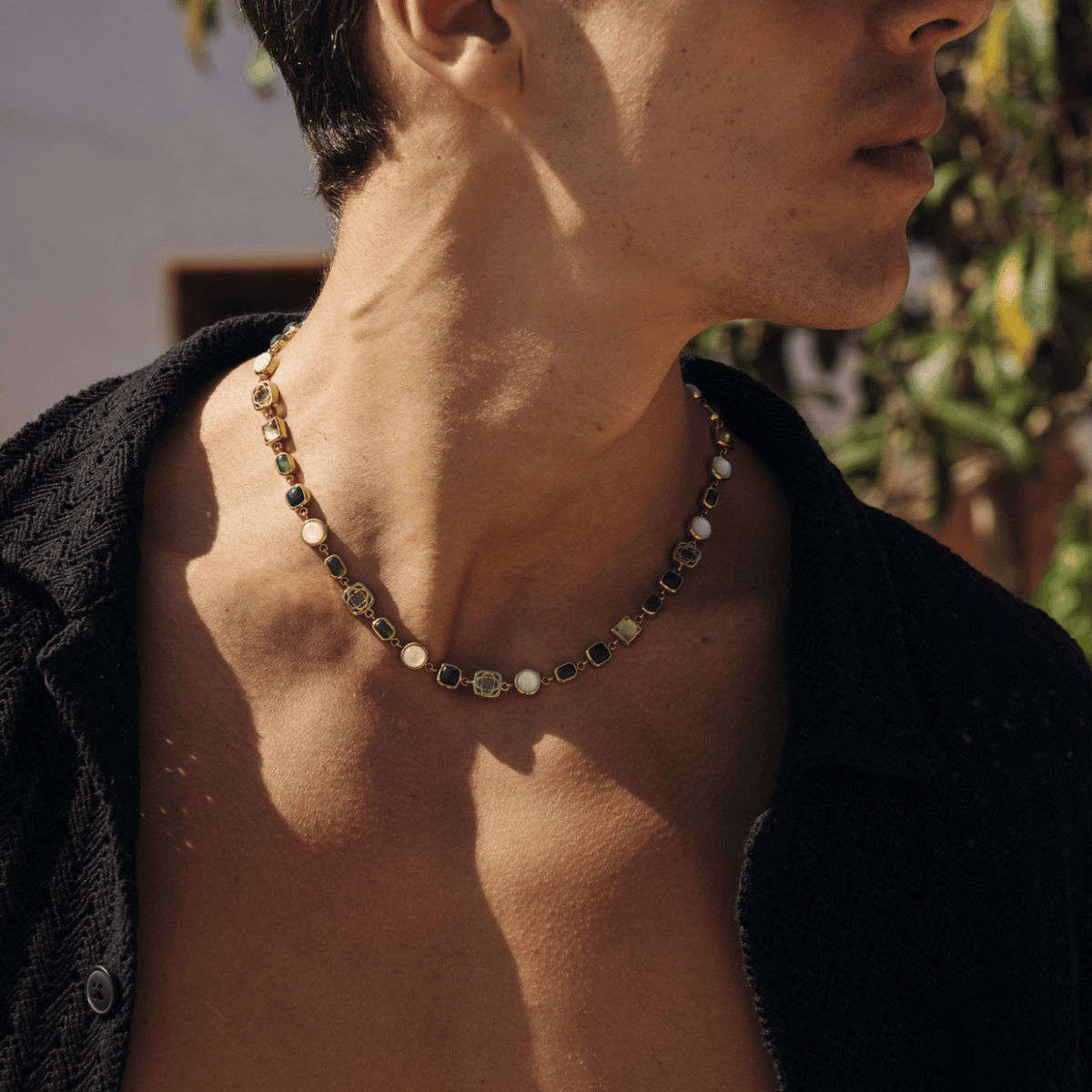 Gold-tone necklace with colored rectangular and pale round accents worn with an open black shirt.