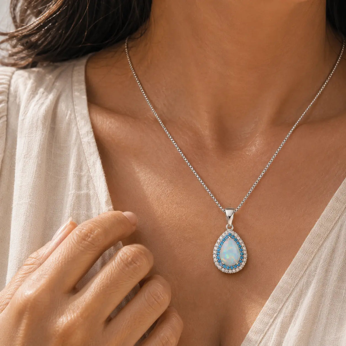 Model wearing a silver-tone necklace with a pale teardrop pendant and blue-and-clear halo.