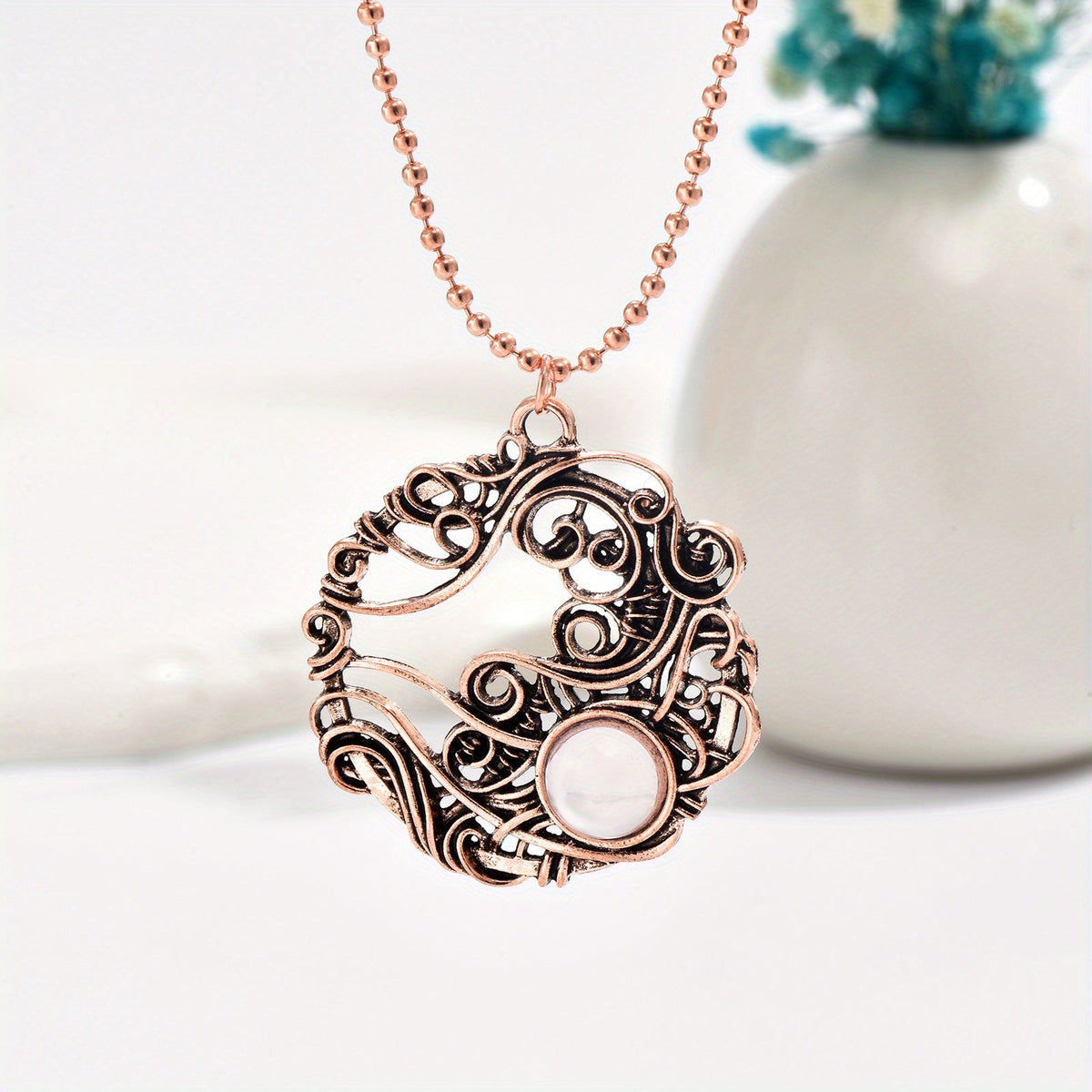 Rose-gold-tone circular wirework pendant with curling details, a pale round accent, and matching chain.