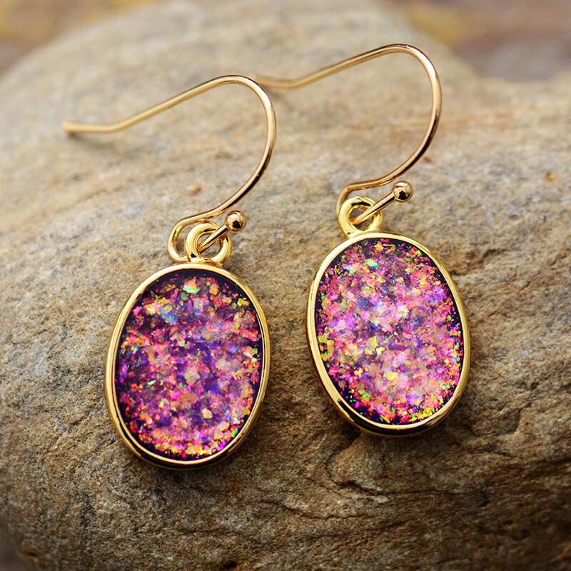 Pink iridescent-looking oval drop earrings with slim gold-tone borders resting on a textured surface.